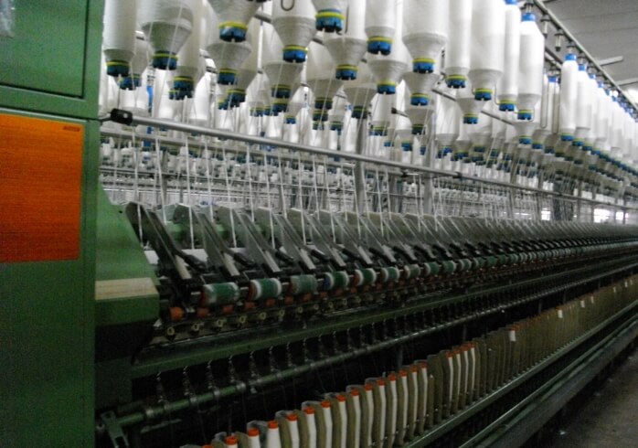 画像:紡績工場の精紡機。上部に並んだ白い糸巻きから糸が引き出され、下のボビンに細く巻き取られていく様子。